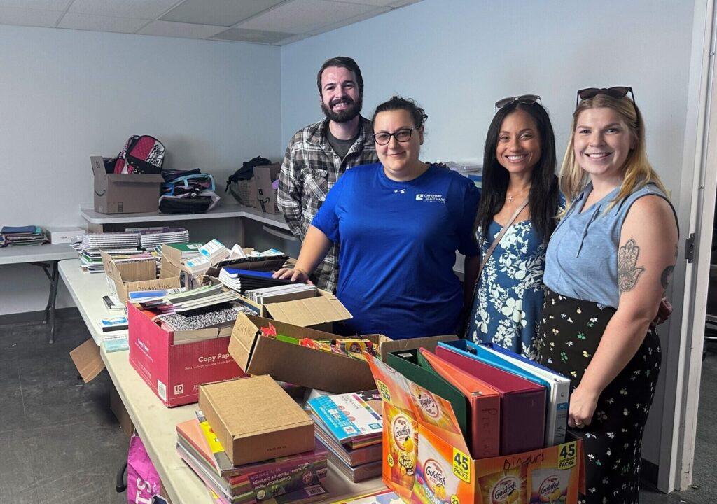 4 smiling Capehart Scatchard attorneys standing behind a table full of school supplies which are being sorted